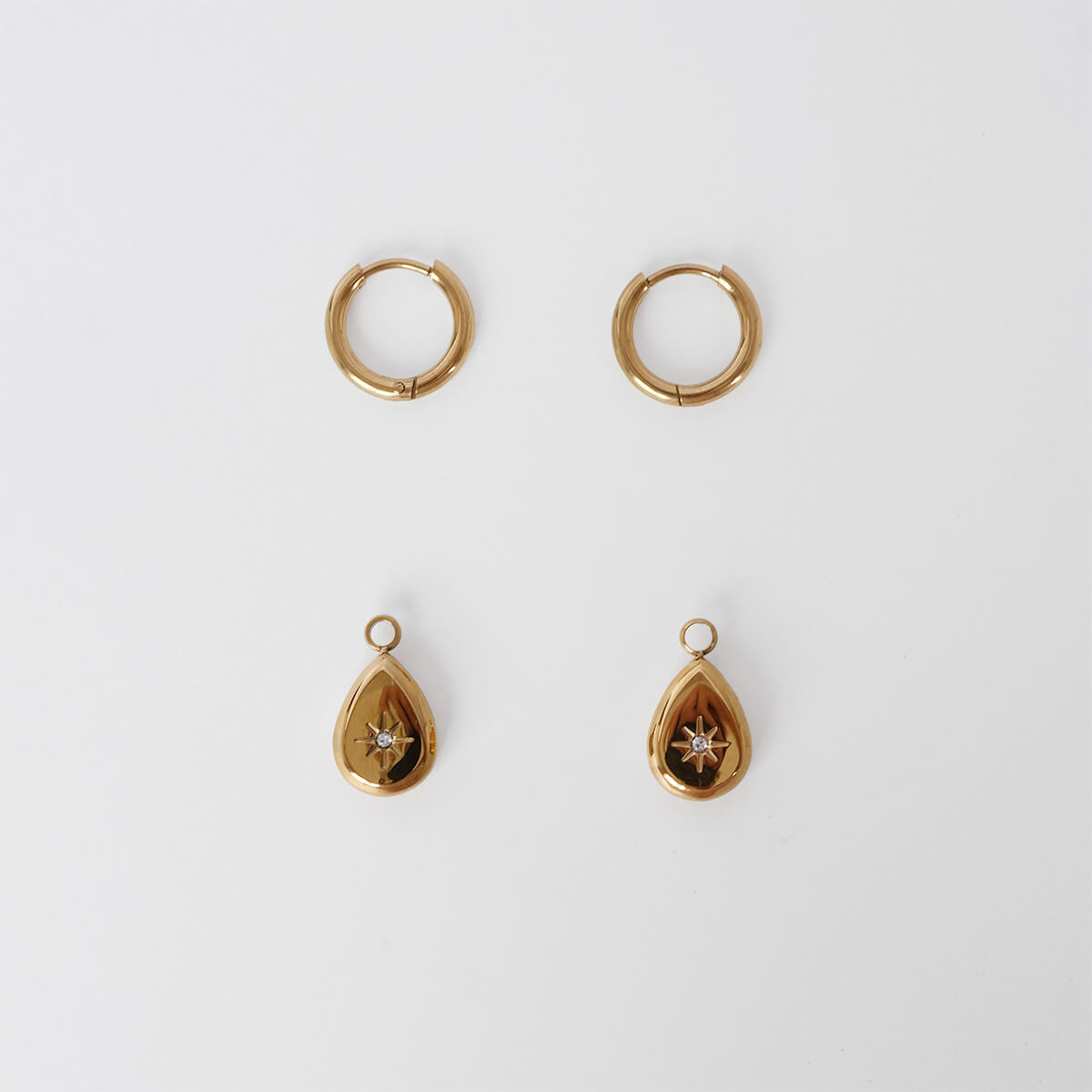 A pair of gold huggie hoop earrings and a pair of gold teardrop pendant earrings with a small star detail, arranged symmetrically on a light gray background.