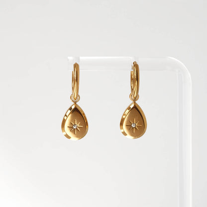A pair of gold huggie hoop earrings with teardrop-shaped dangling pendants, each featuring a small starburst detail and a tiny clear gemstone, displayed on a clear acrylic stand against a white background.