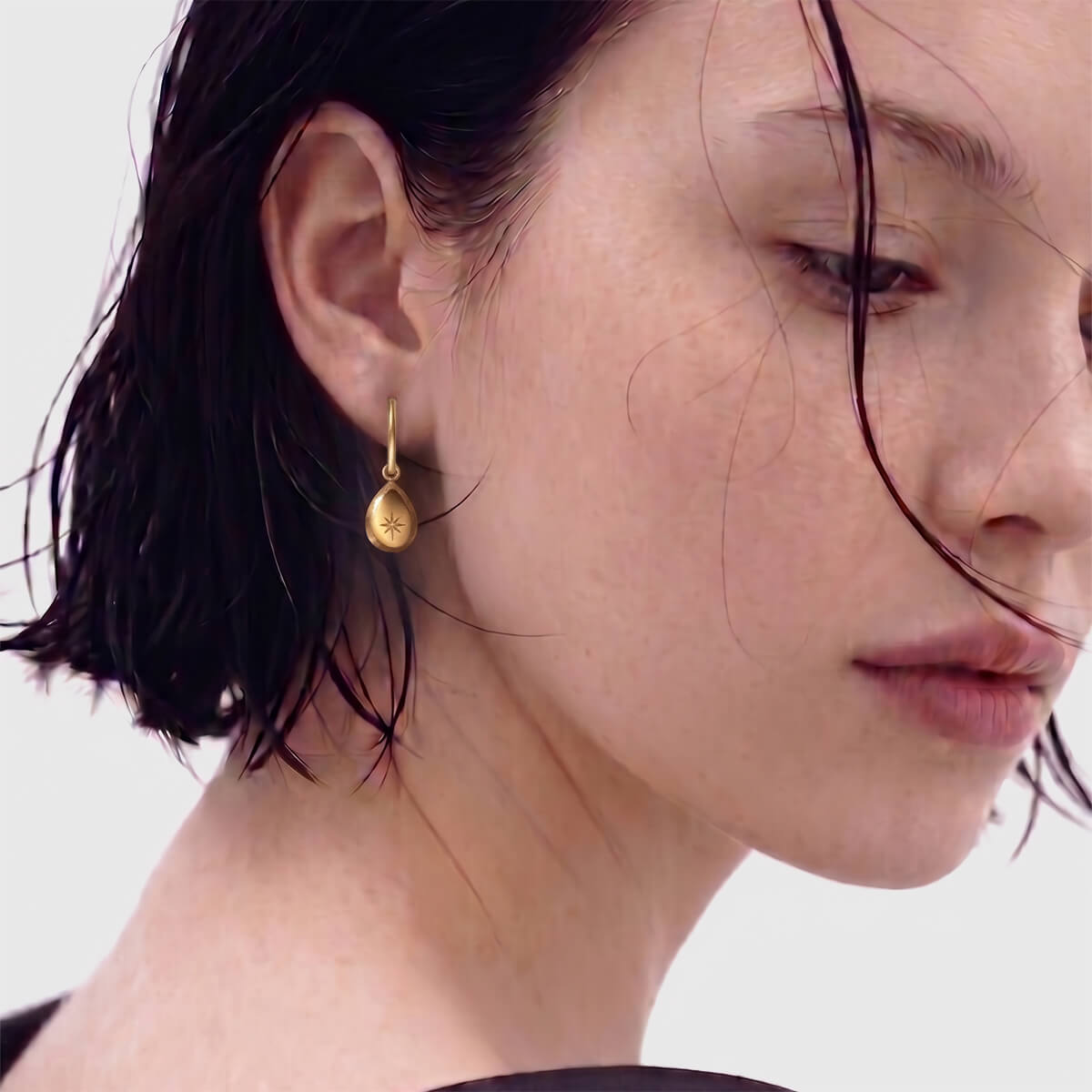 A close-up of a model wearing a small gold teardrop dangle earring against a light gray background.