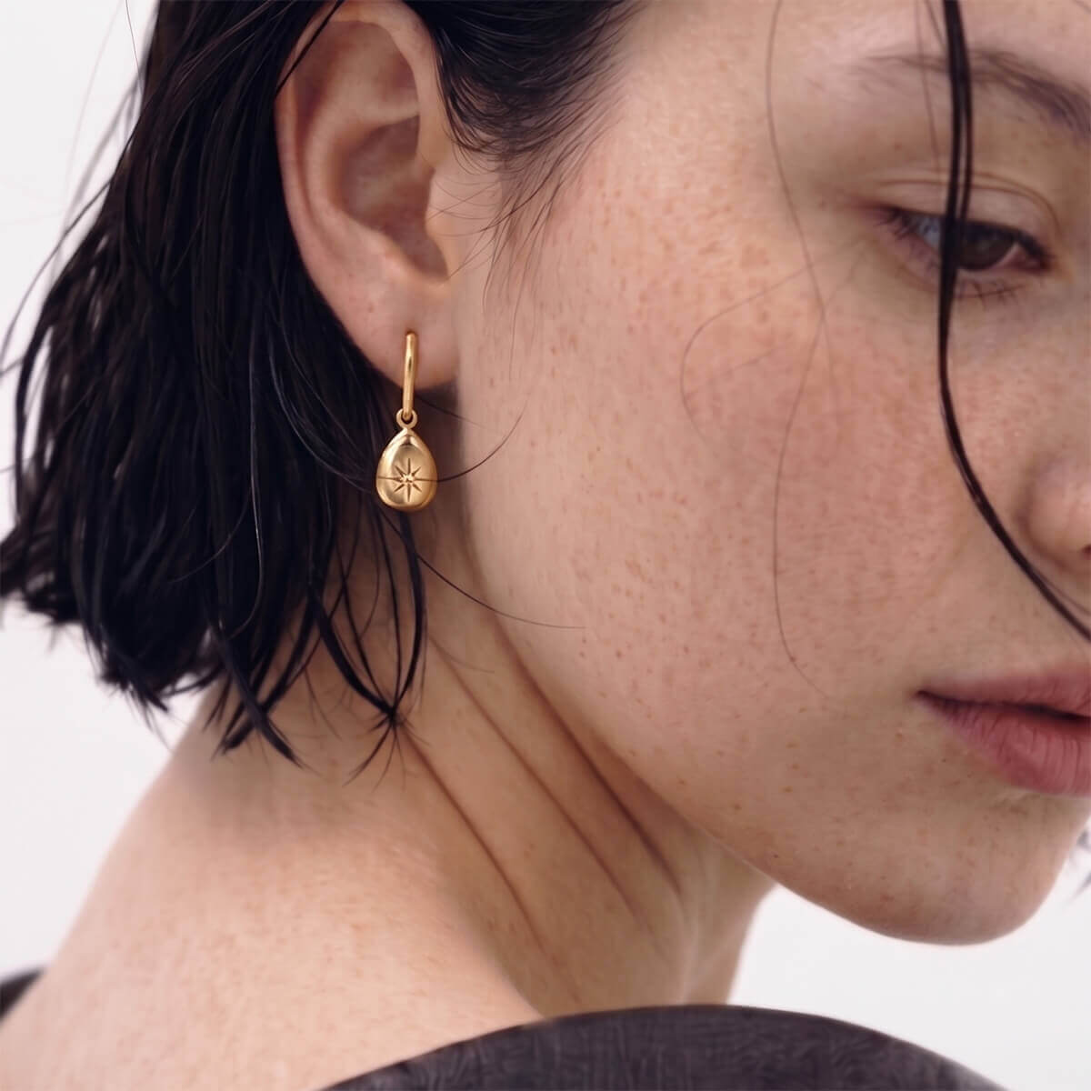 A close-up of a model's ear wearing a small gold teardrop dangle earring with a starburst pattern, set against a clean white background.