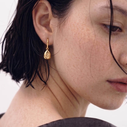 A close-up of a model's ear wearing a small gold teardrop dangle earring with a starburst pattern, set against a clean white background.