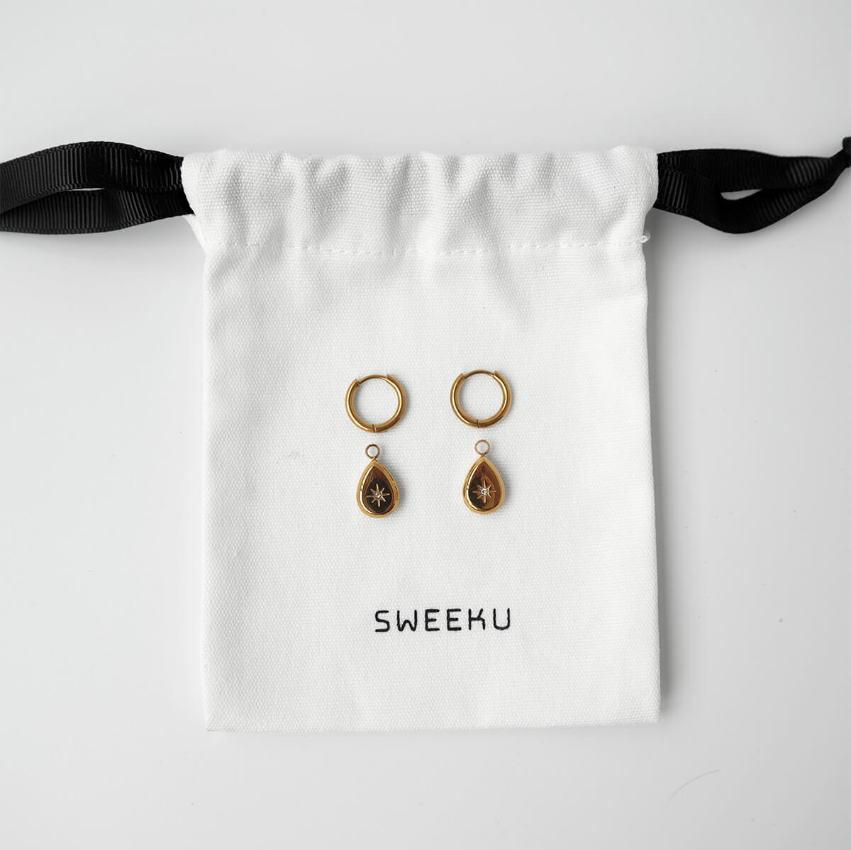 A pair of gold-toned dangle earrings with teardrop-shaped drops featuring a star motif, displayed on a white drawstring pouch with the brand name 'Sweeku' printed on it.