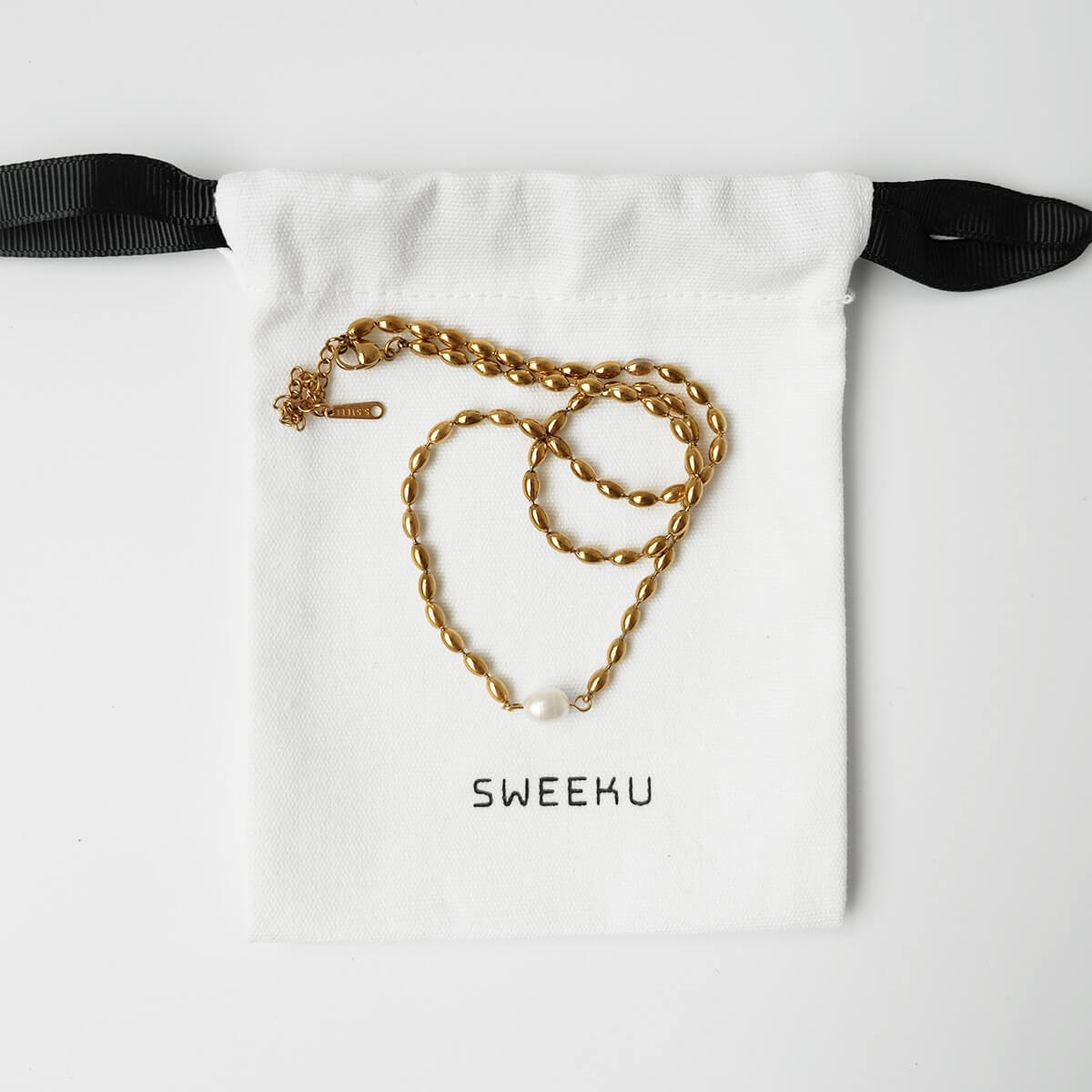 A gold chain necklace with a small white pearl pendant is displayed on a white drawstring pouch with black ribbon ties. The pouch is printed with the brand name 'SWEEKU' in black text.