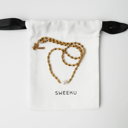 A gold chain necklace with a small white pearl pendant is displayed on a white drawstring pouch with black ribbon ties. The pouch is printed with the brand name 'SWEEKU' in black text.